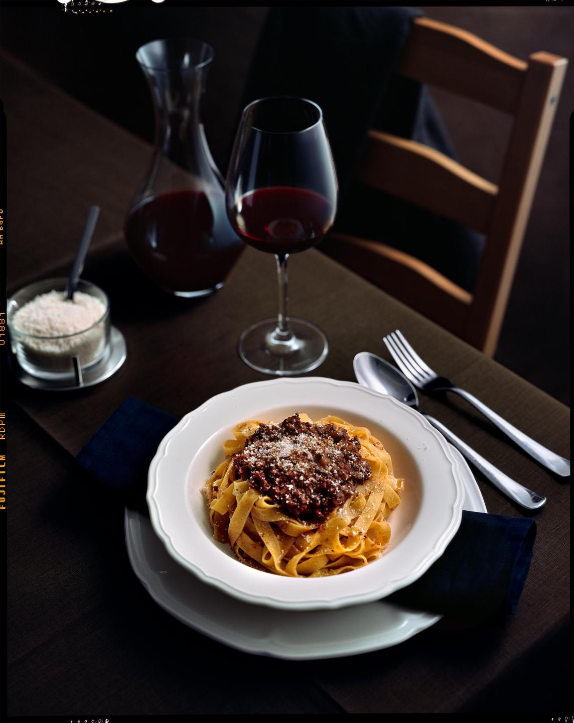 A bowl of tagliatelle bolognese with a glass of red wine and a decanter are arranged on a table.