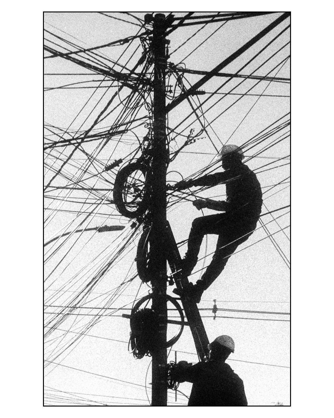 Two men are working on a utility pole with many wires against a bright sky.
