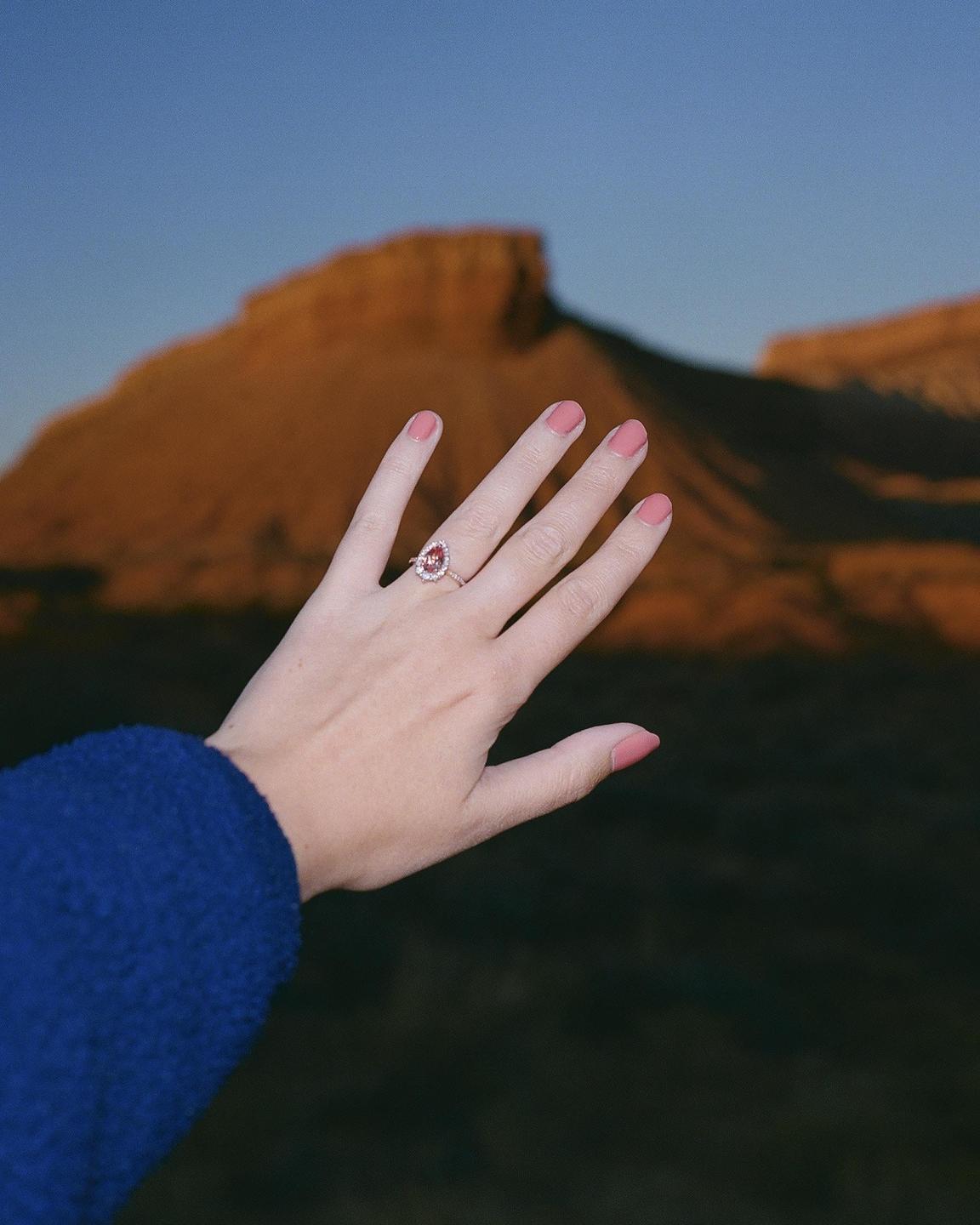 A hand with a ring and pink nail polish is held up against a desert mountain landscape at dusk.