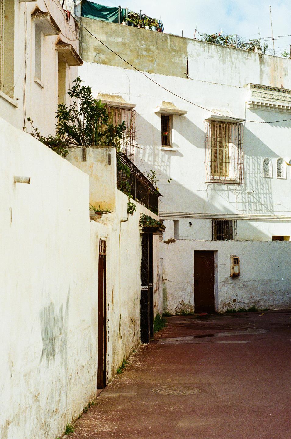 A narrow street with white buildings and dark wooden doors is bathed in sunlight, casting shadows on the walls.