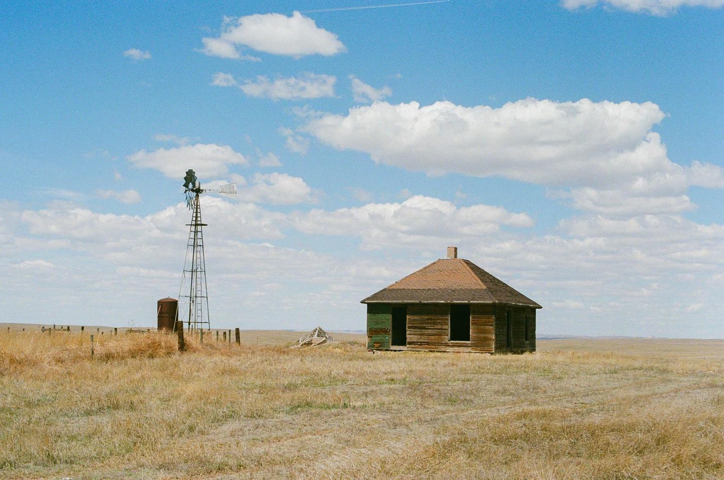 An abandoned wooden house with a windmill stands in a vast field under a blue sky with clouds.