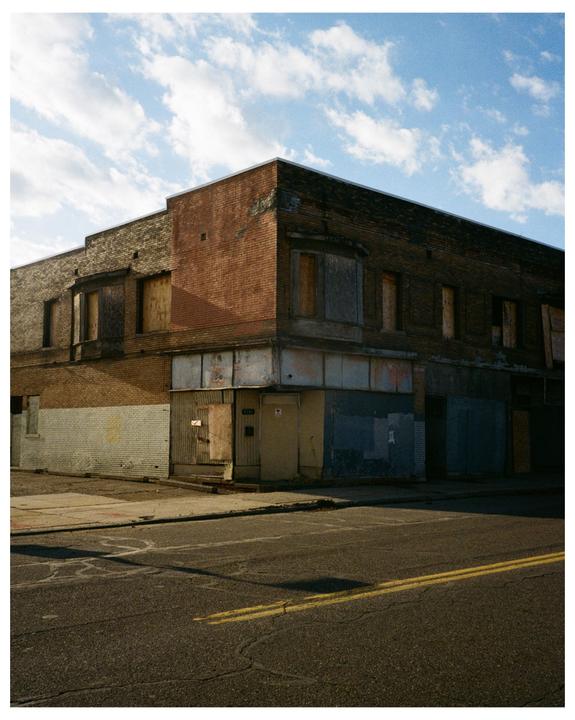 An abandoned brick building with boarded-up windows stands at a street corner under a cloudy sky.