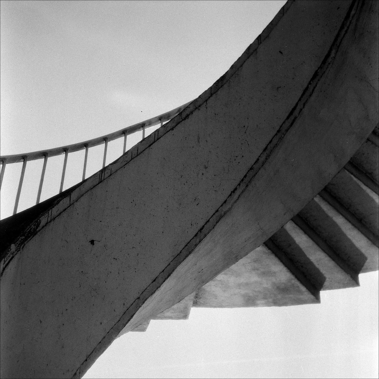 A concrete spiral staircase with a metal railing ascends towards a bright sky, casting shadows on its underside.