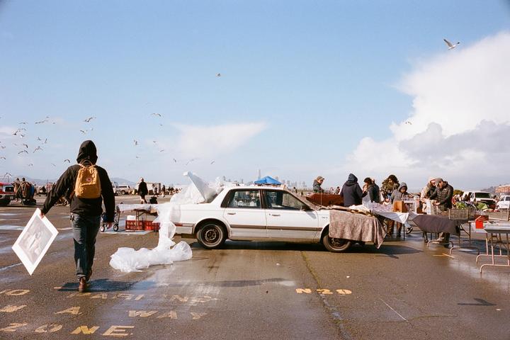 A person with a backpack walks away from a white car and market stalls with people browsing.