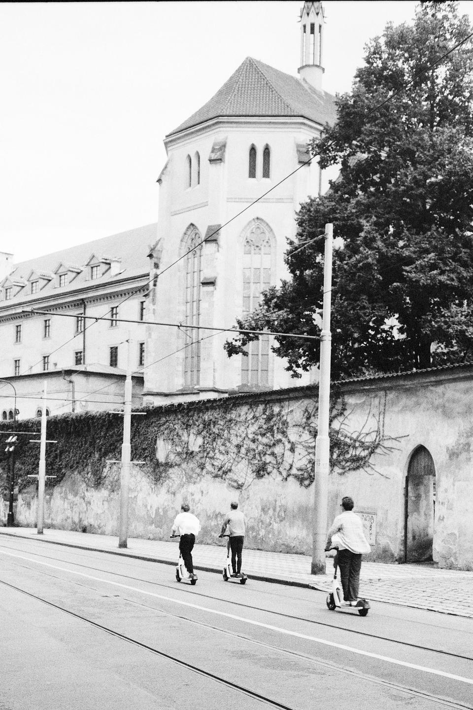 Three men ride electric scooters down a street next to a stone wall covered in ivy and a large building with a tall, pointed roof.