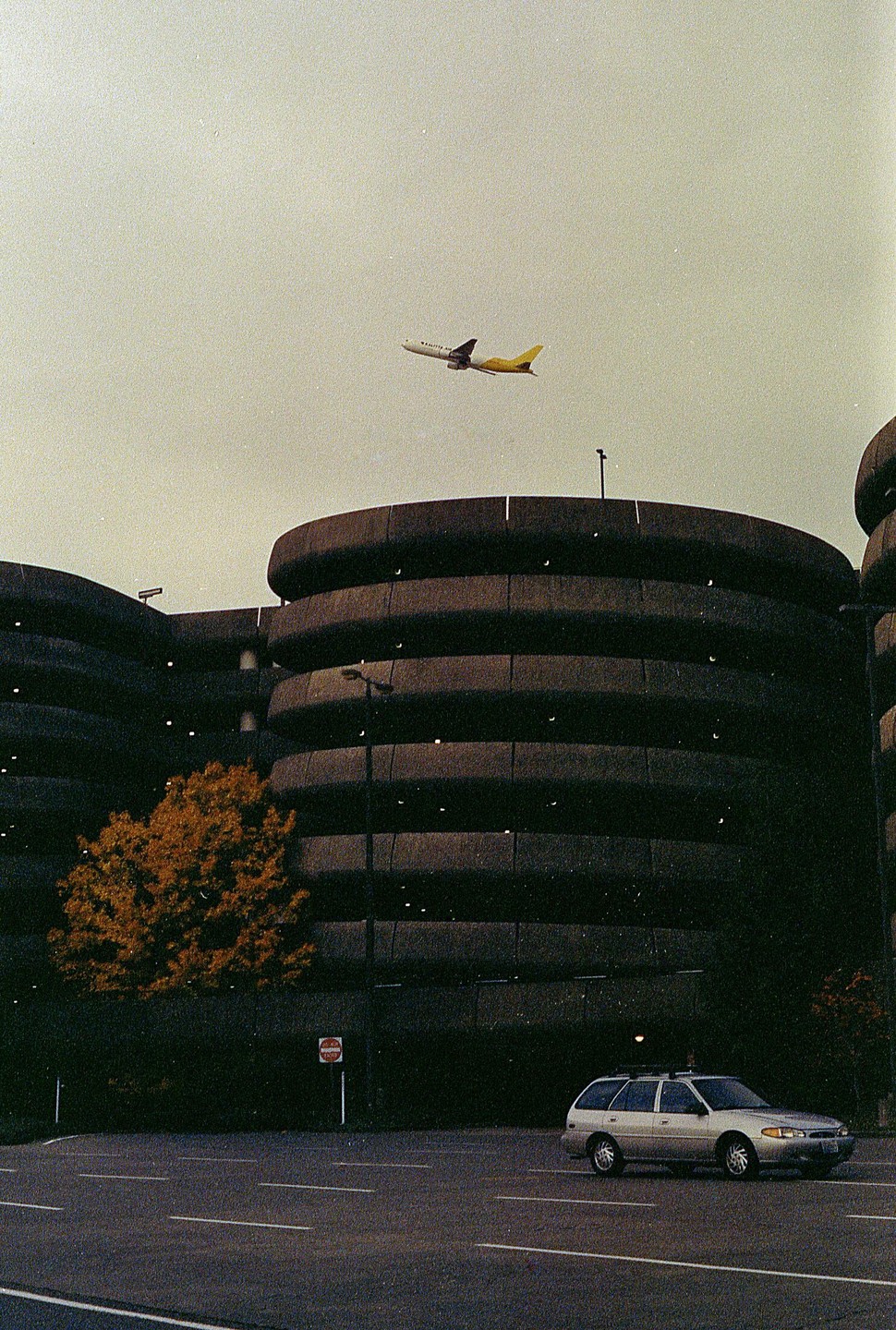 An airplane flies over a circular parking garage with a car parked in front.