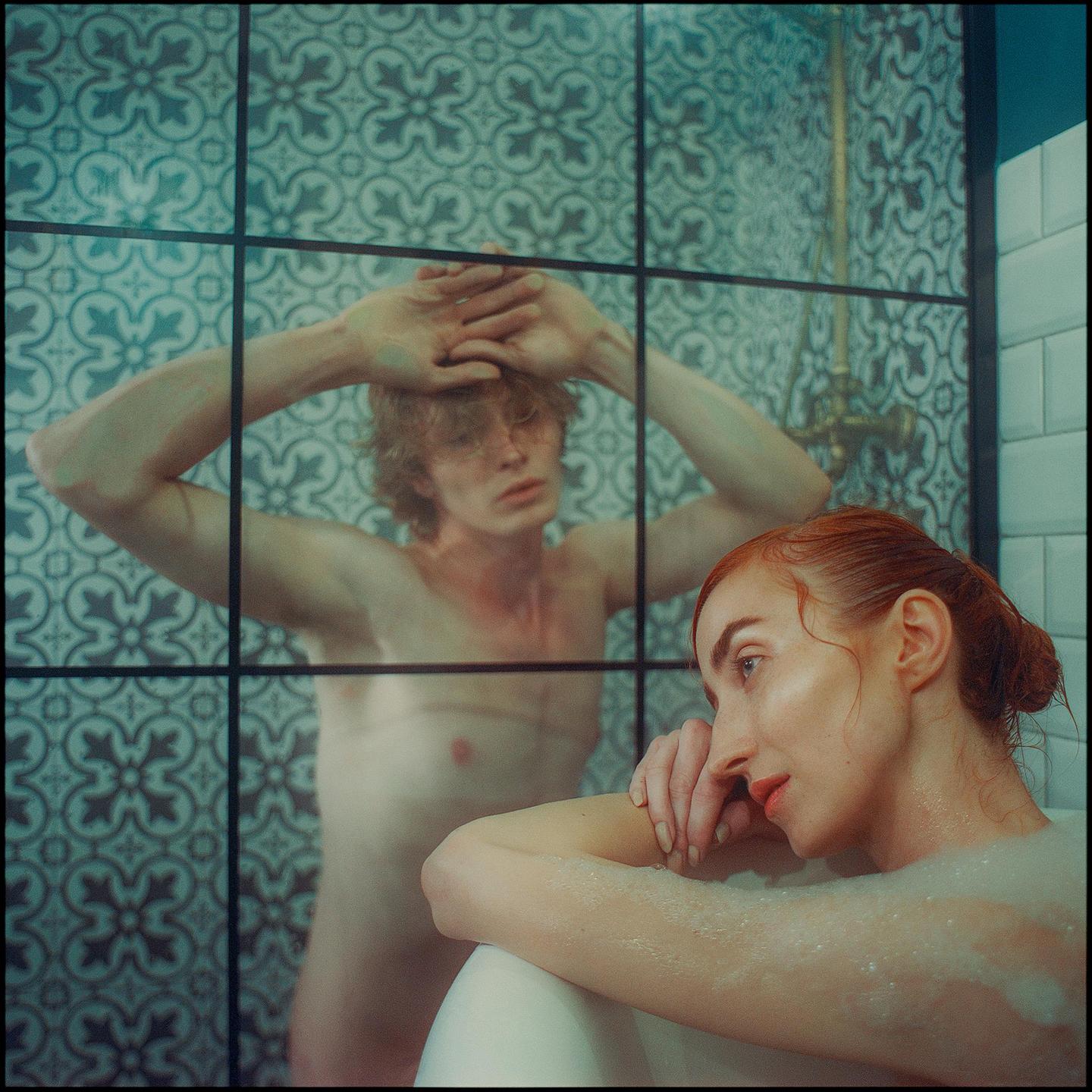 A woman in a bathtub looks away from a man standing behind a glass shower door.