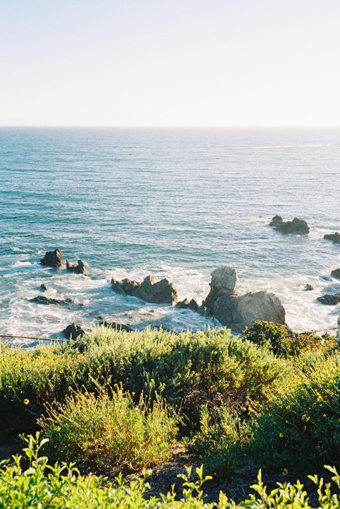 The Pacific Ocean with rocky outcrops and green vegetation in the foreground is visible under a clear sky.