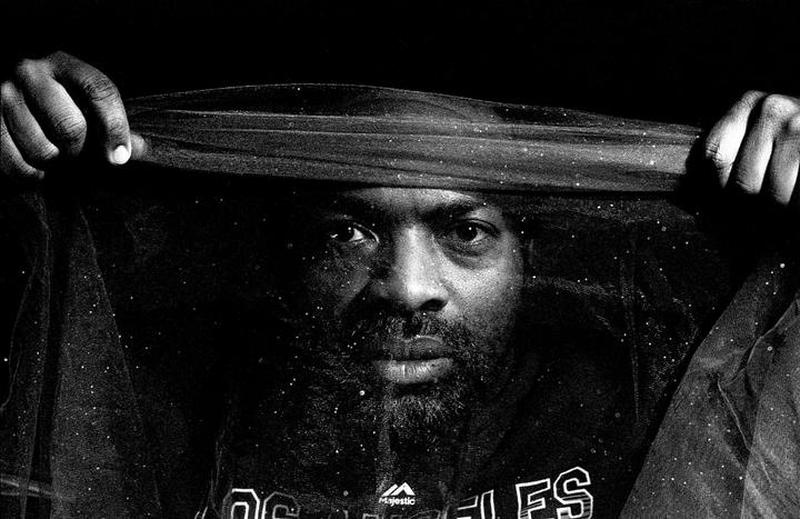 A man with a beard holds a sheer, glittery fabric over his head, looking directly at the viewer.