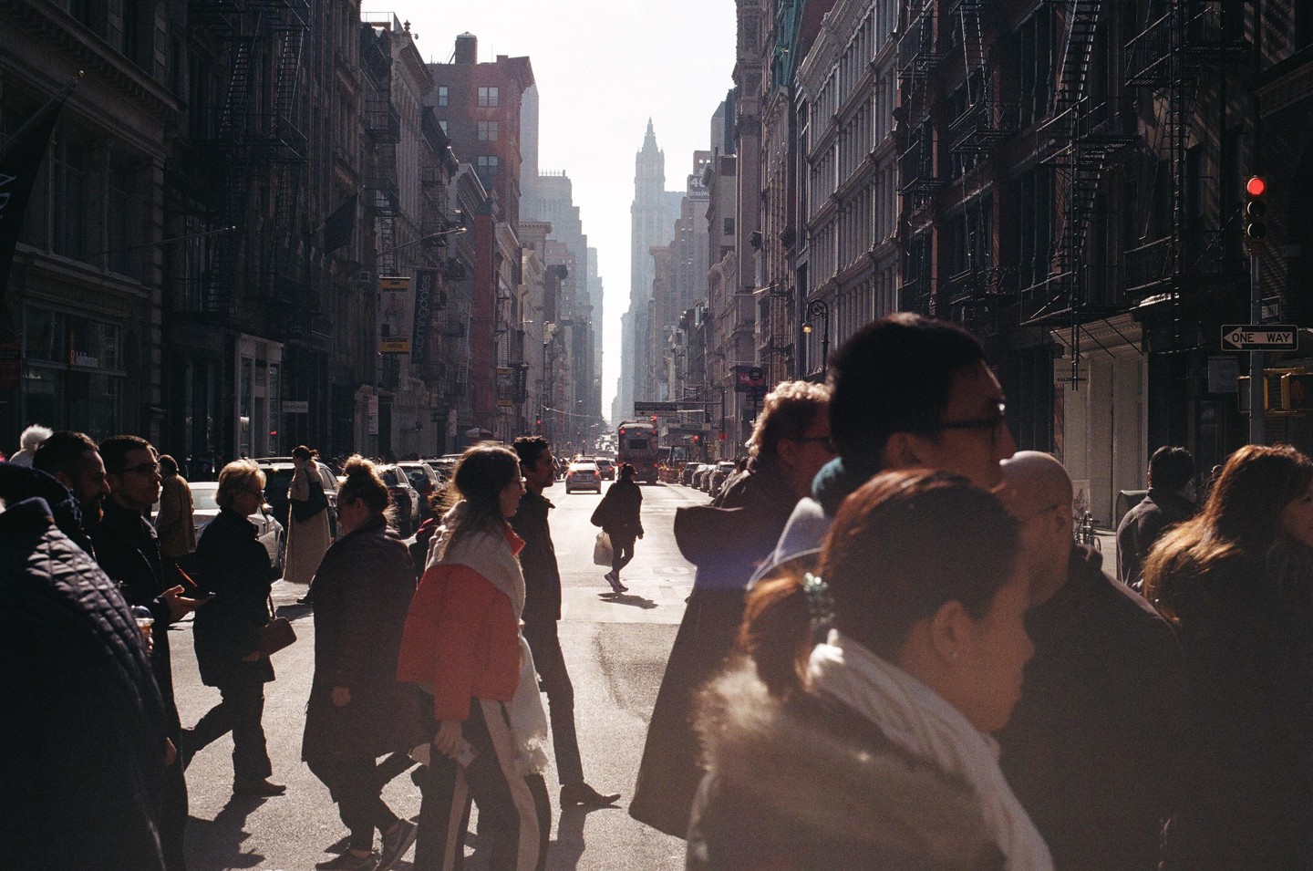 A busy street scene shows many pedestrians crossing a sunlit intersection with tall buildings lining both sides of the road.