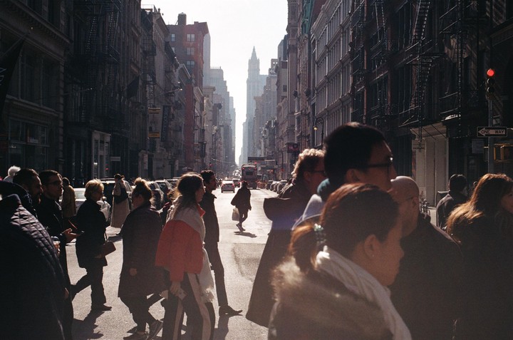 A busy street scene shows many pedestrians crossing a sunlit intersection with tall buildings lining both sides of the road.