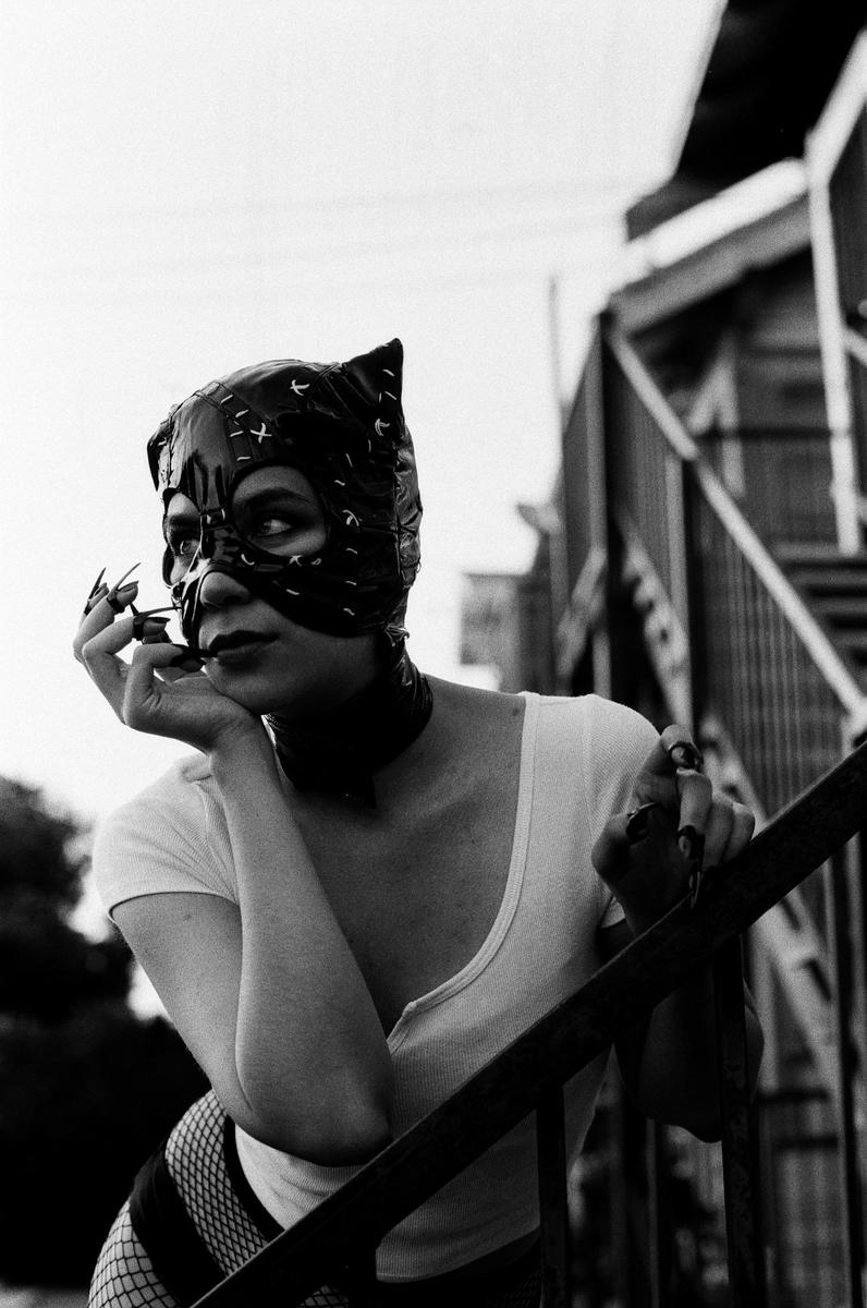 A woman in a cat mask and fishnet stockings leans on a railing outdoors.