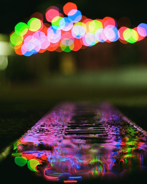 Colorful bokeh lights are reflected in a wet surface, possibly a street or sidewalk, at night.