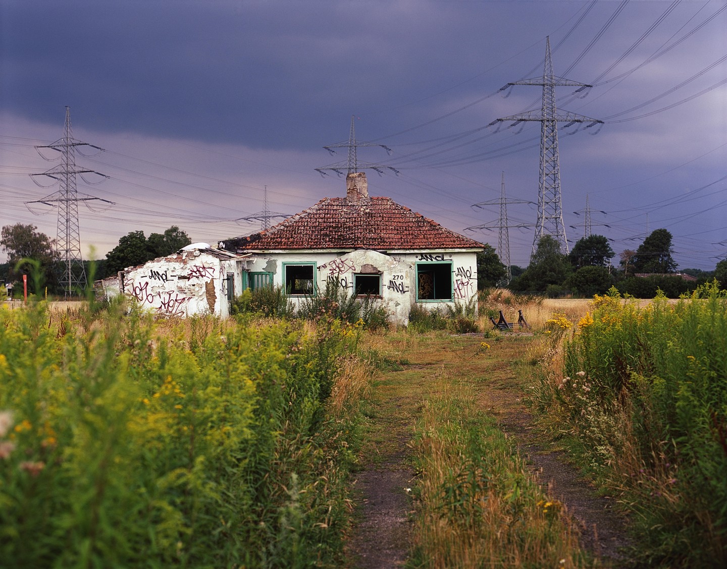 An abandoned house with a red tile roof and graffiti on its walls stands in a field with tall grass and power lines under a dark sky.