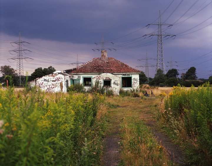 An abandoned house with a red tile roof and graffiti on its walls stands in a field with tall grass and power lines under a dark sky.