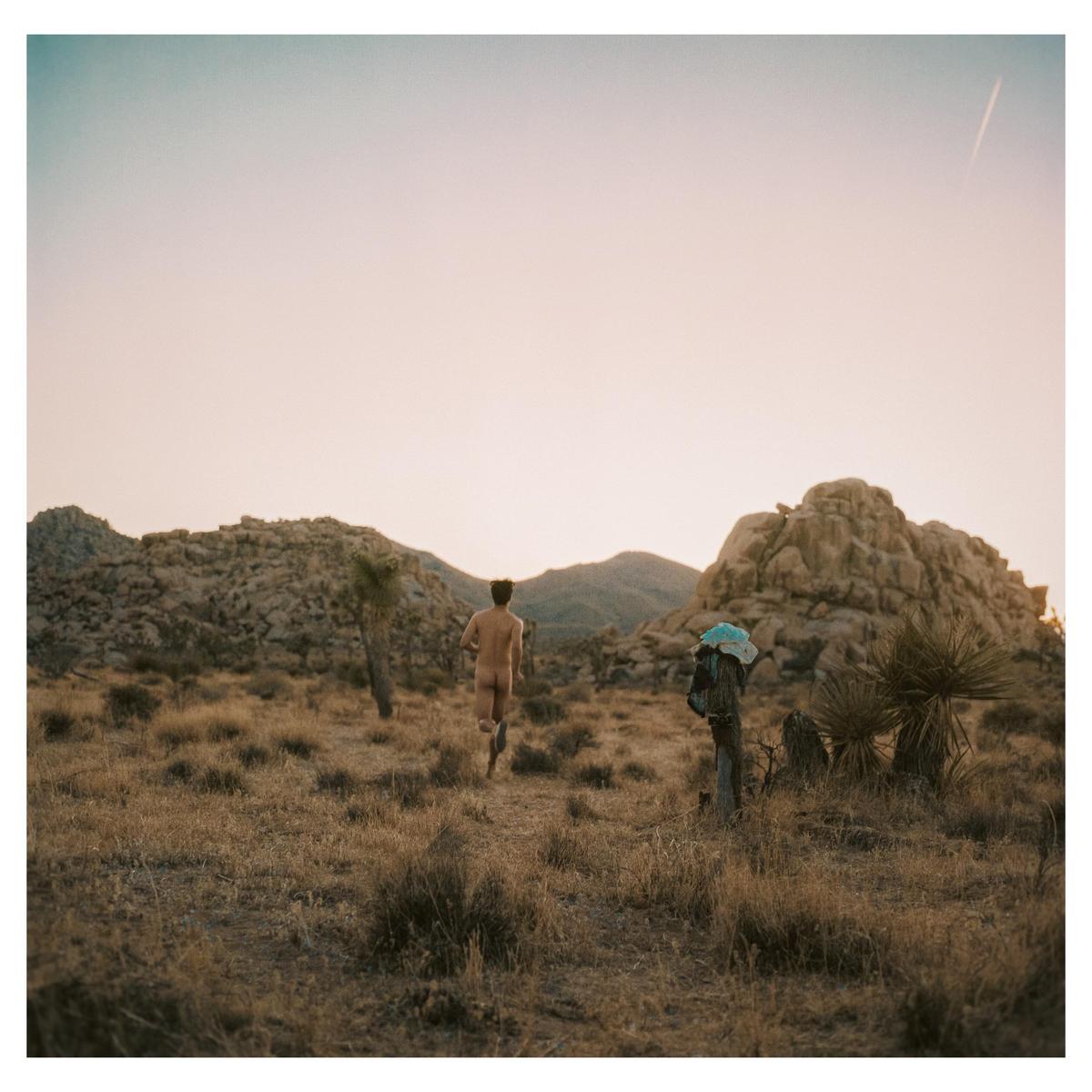 A man runs naked through a desert landscape with mountains and sparse vegetation under a clear sky.