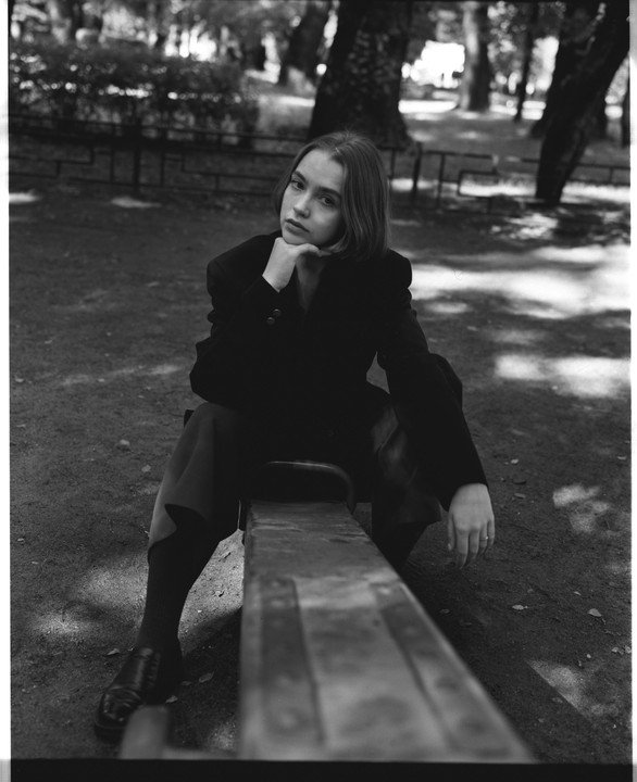 A woman in a blazer sits on a park bench with her chin resting on her hand.