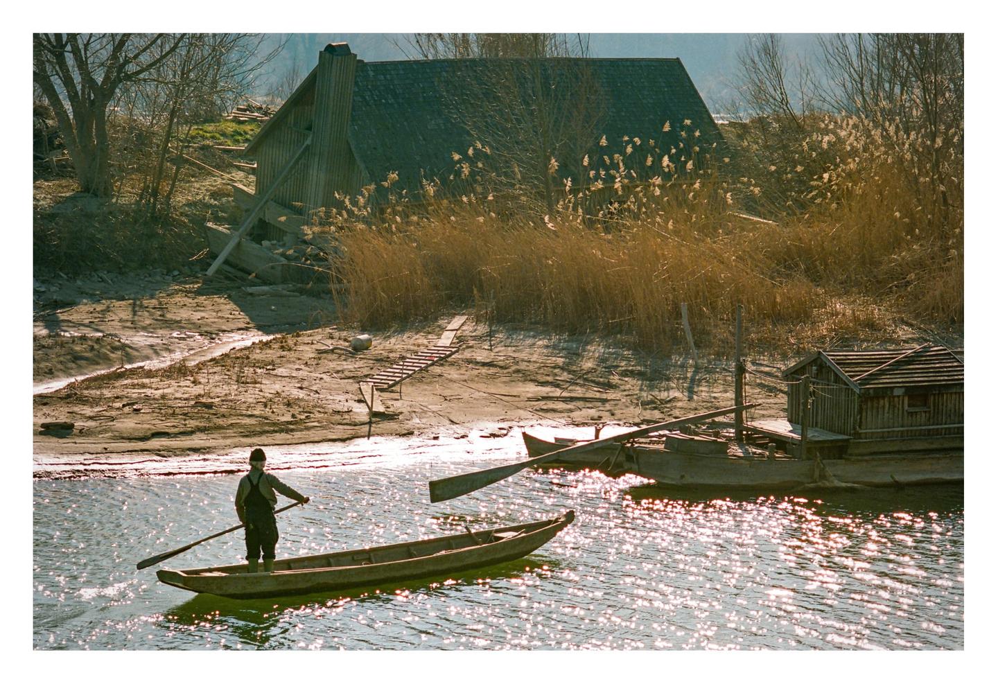 A man stands in a small boat on sparkling water, with a wooden shack and tall dry grass on the shore behind him.