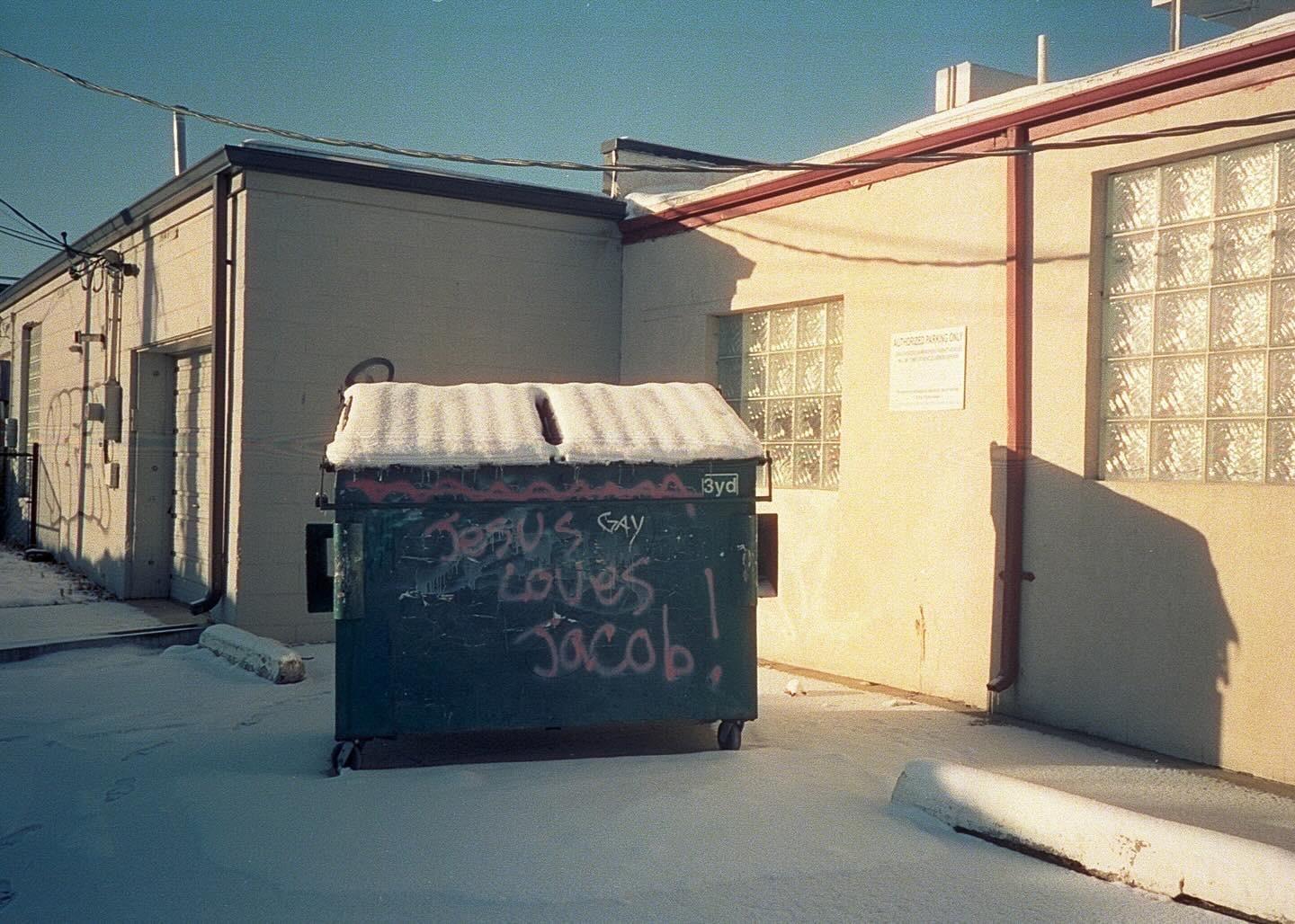 A snow-covered dumpster with graffiti stands in an alleyway between two buildings.