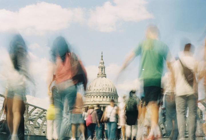 A blurred crowd of people walks on a bridge with St. Paul's Cathedral visible in the background.