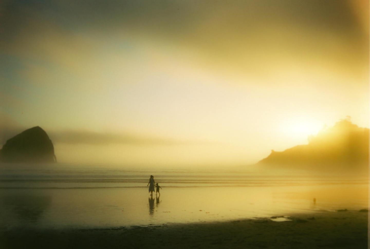 A woman and child walk along a misty beach at sunset with a large rock formation in the background.