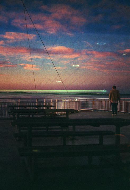 A man stands on a ferry deck looking out at the ocean under a colorful sunrise sky.