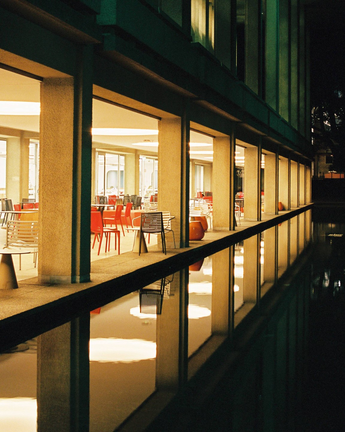 A building with many windows and pillars is reflected in a body of water at night.