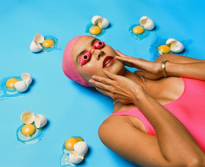 A woman in a pink swimsuit and swim cap lies on a blue surface surrounded by broken eggs.