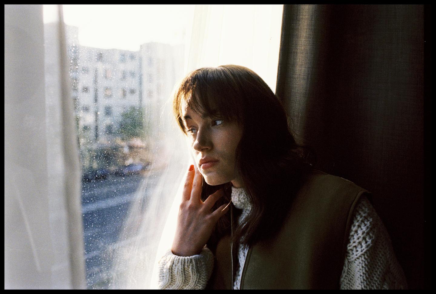 A woman with long hair and bangs looks out a window with her hand to her face.