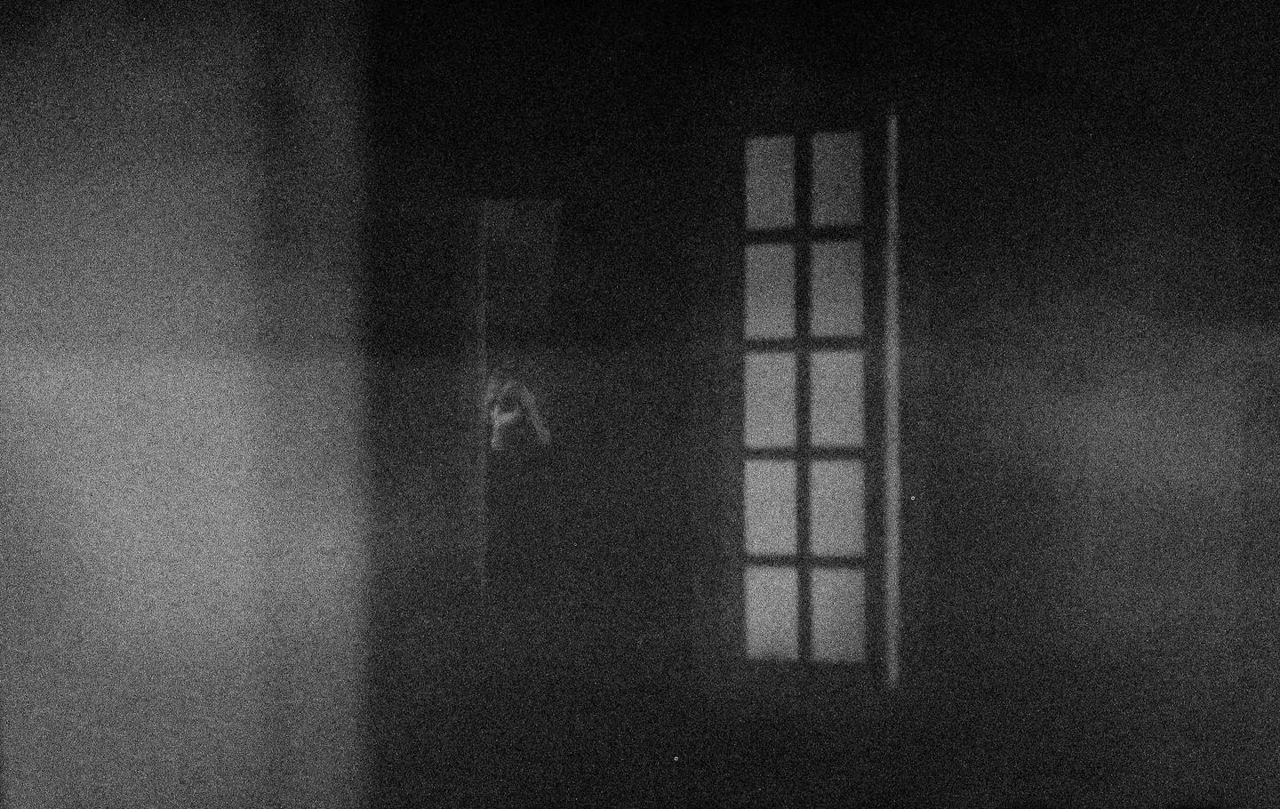 A dark room with a window showing a grid pattern and a faint, blurry shape of a person.
