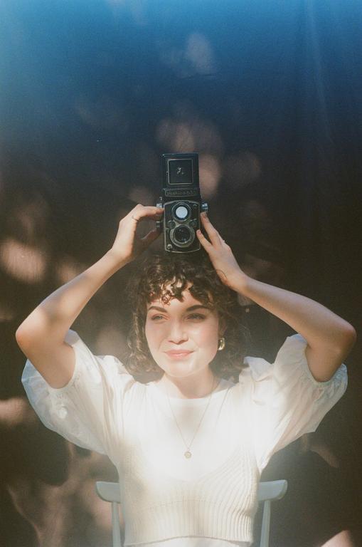 A woman with curly hair holds a twin-lens reflex camera above her head, smiling at the viewer.