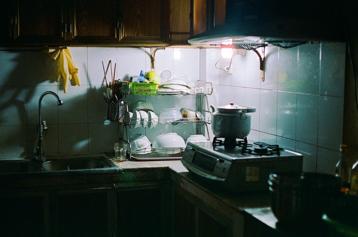 A kitchen counter with a sink, a dish rack, and a stove with a pot on it is illuminated by a single light.