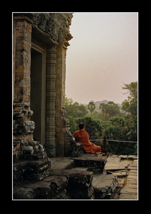 A monk in an orange robe sits on stone steps looking out at a lush green landscape with palm trees.