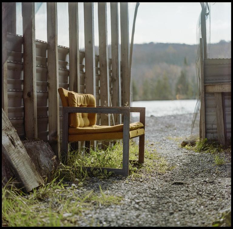 A yellow armchair sits on a gravel path next to a wooden fence, with a lake and forest in the background.