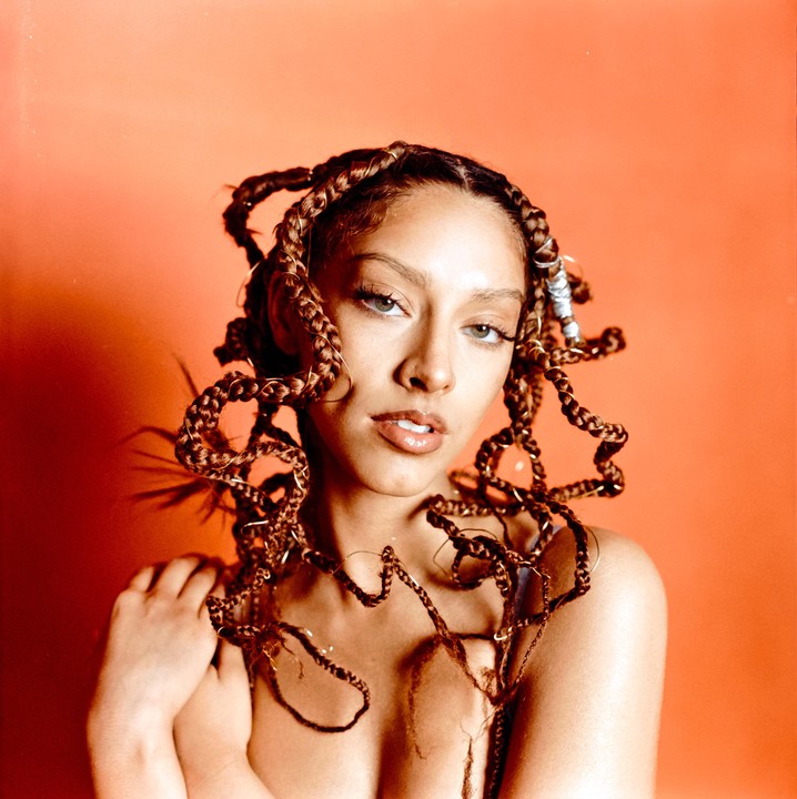A woman with braided hair and rings in her braids poses against an orange background.