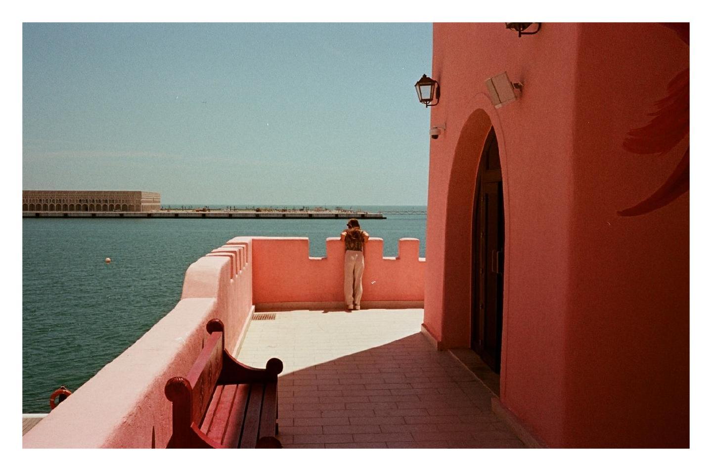 A person stands on a pink balcony overlooking a body of water with a building in the distance.