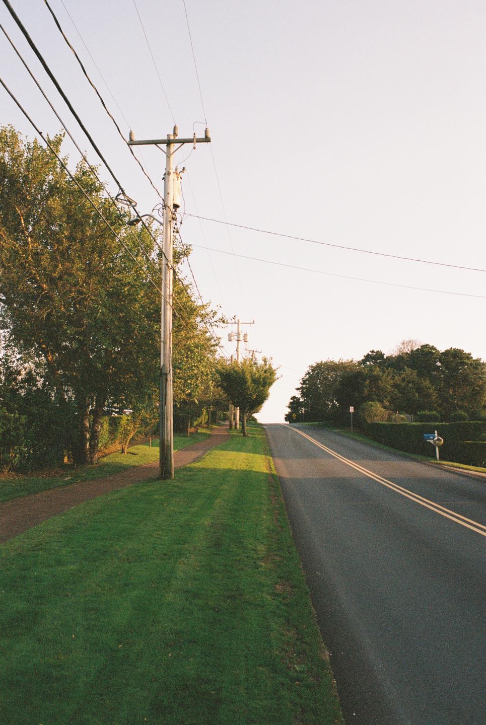 A road with a sidewalk and power lines runs uphill between trees and grass.