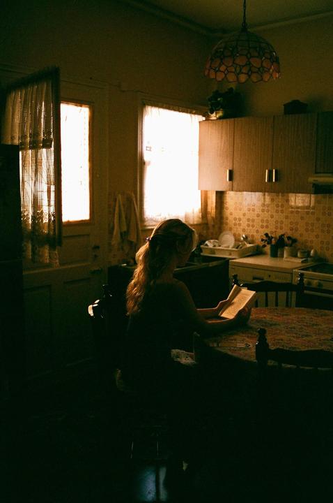 A woman with long hair sits at a table in a dimly lit kitchen, reading a book.