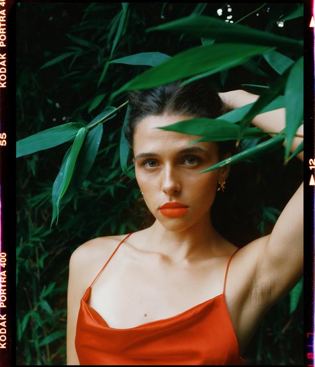 A woman in a red dress with red lipstick poses among green foliage.