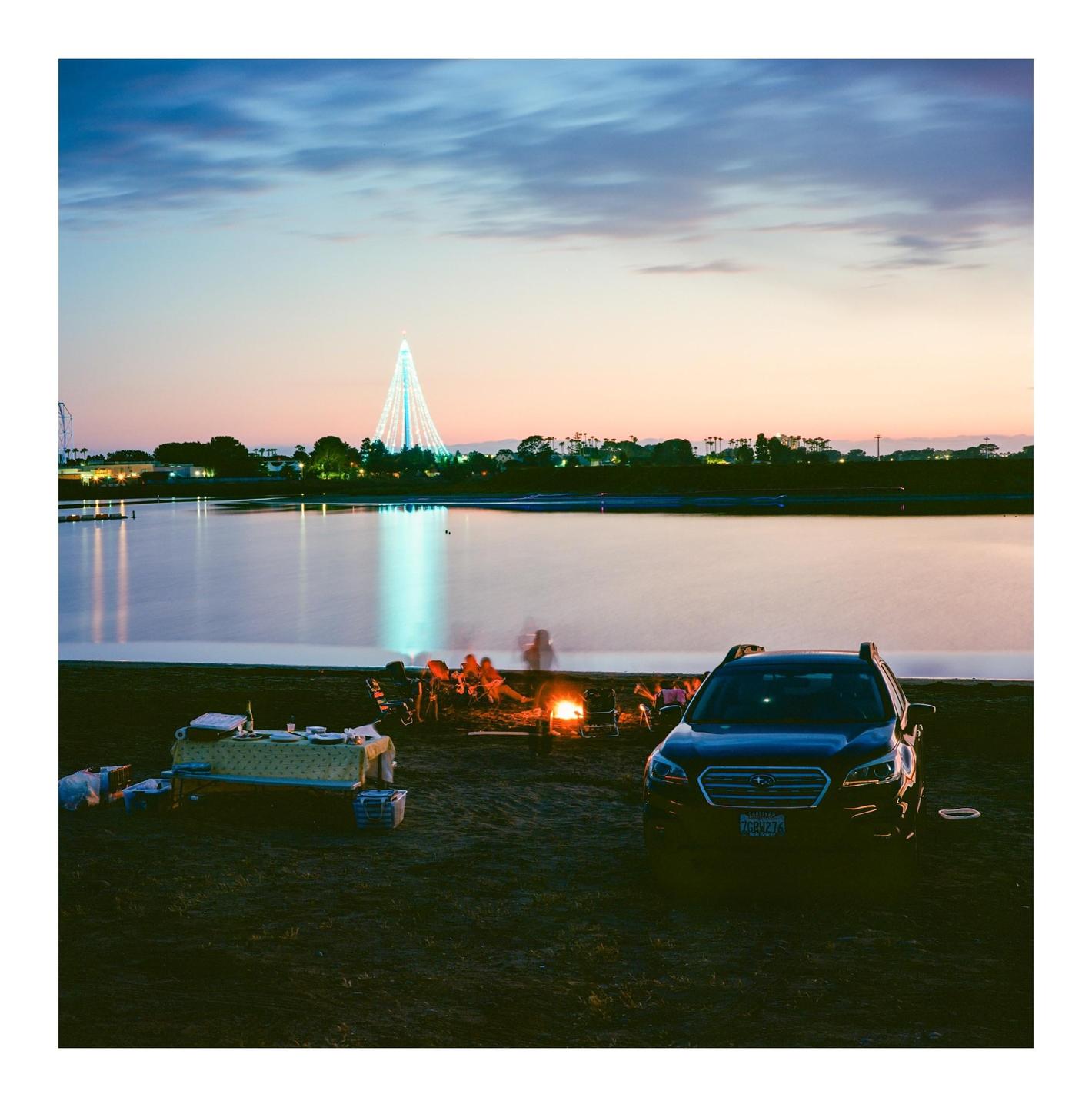 A group of people sit around a bonfire on a beach next to a lake with a large illuminated structure in the distance at dusk.