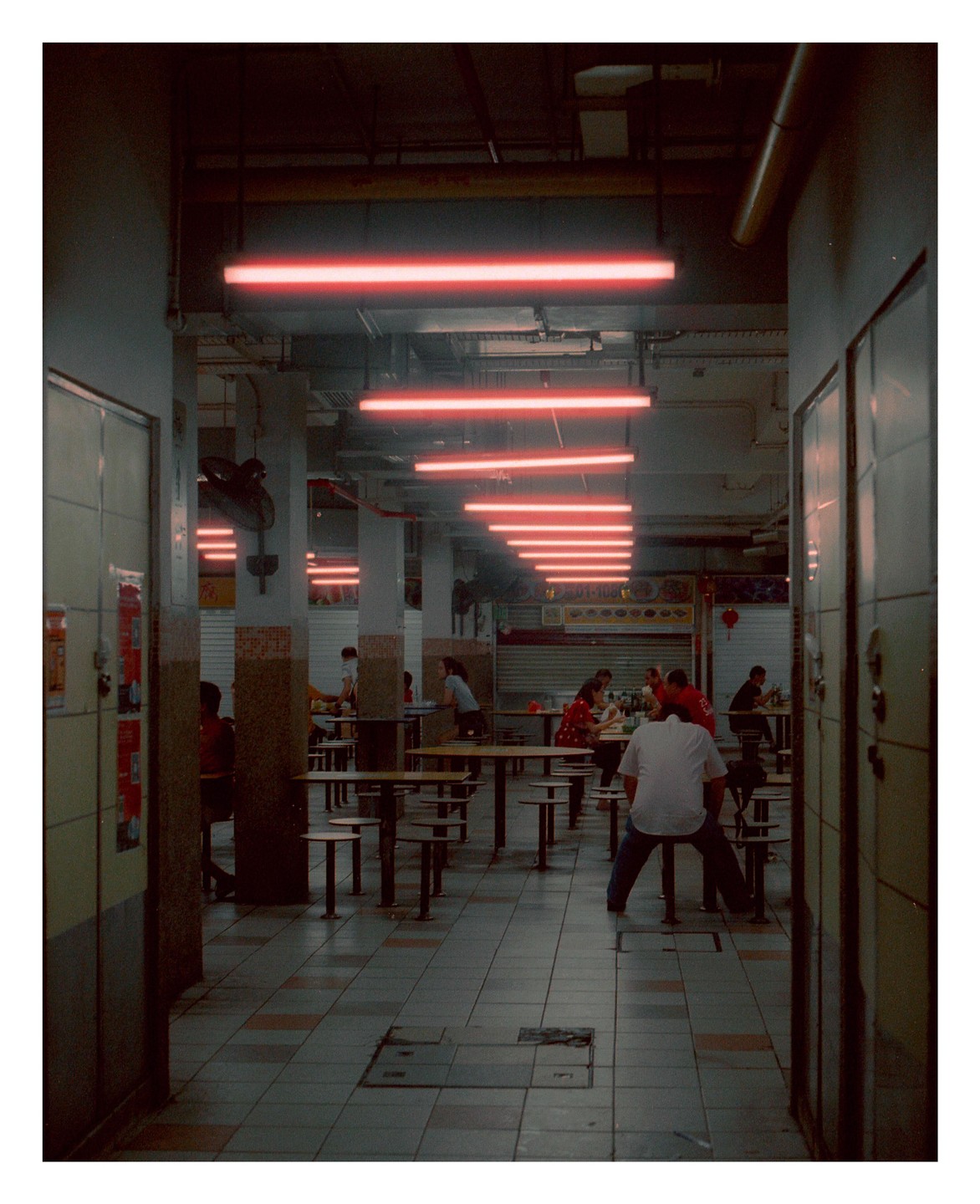 A man sits on a stool in a large, mostly empty food hall lit by red neon lights.