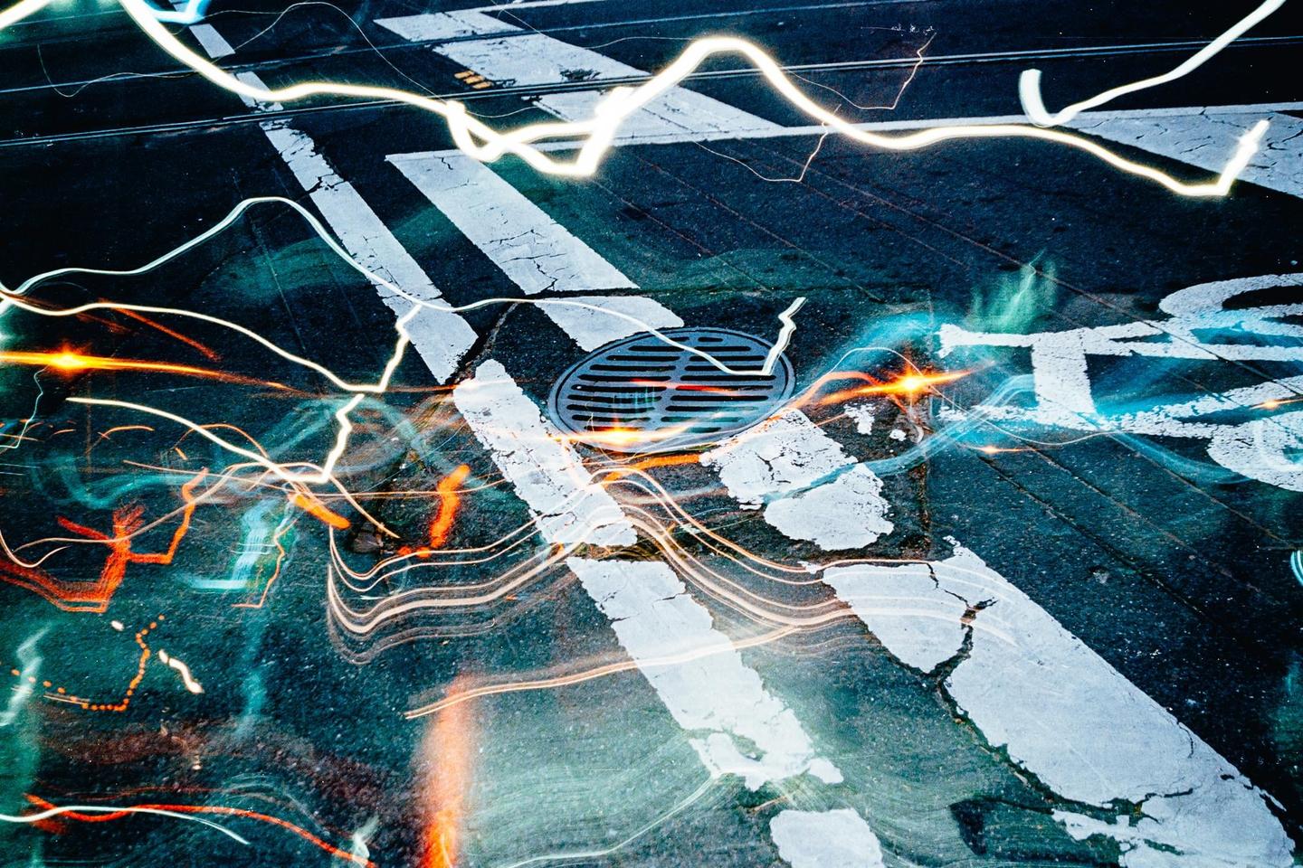 Light trails from vehicles illuminate a street with a manhole cover and painted lines.