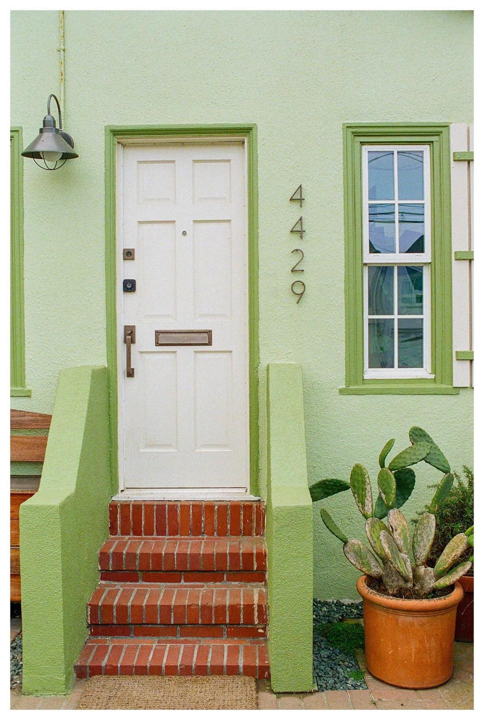 A light green house with a white door and window has brick steps leading up to the entrance, with a potted cactus plant to the right.