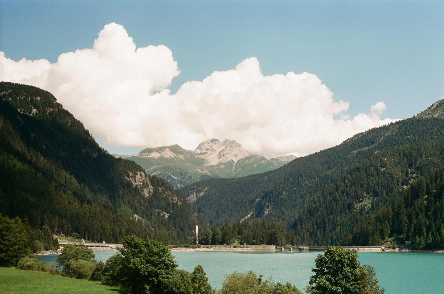 A turquoise lake is nestled between forested mountains under a bright sky with large white clouds.