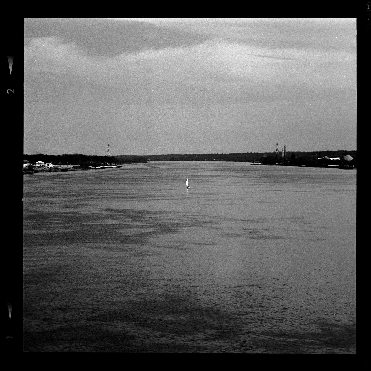 A small sailboat floats in the middle of a wide river with industrial buildings and trees on both banks under a cloudy sky.