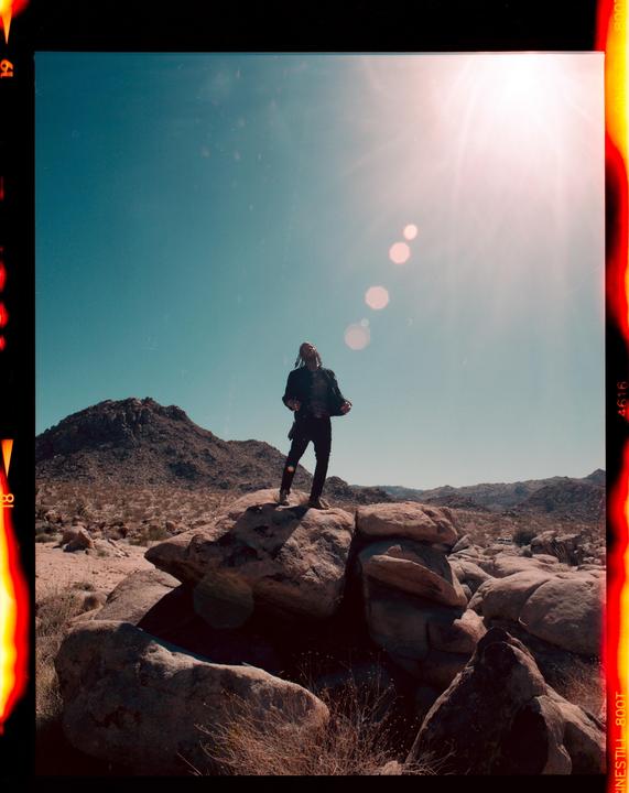 A man stands on a large rock in a desert landscape under a bright sun.