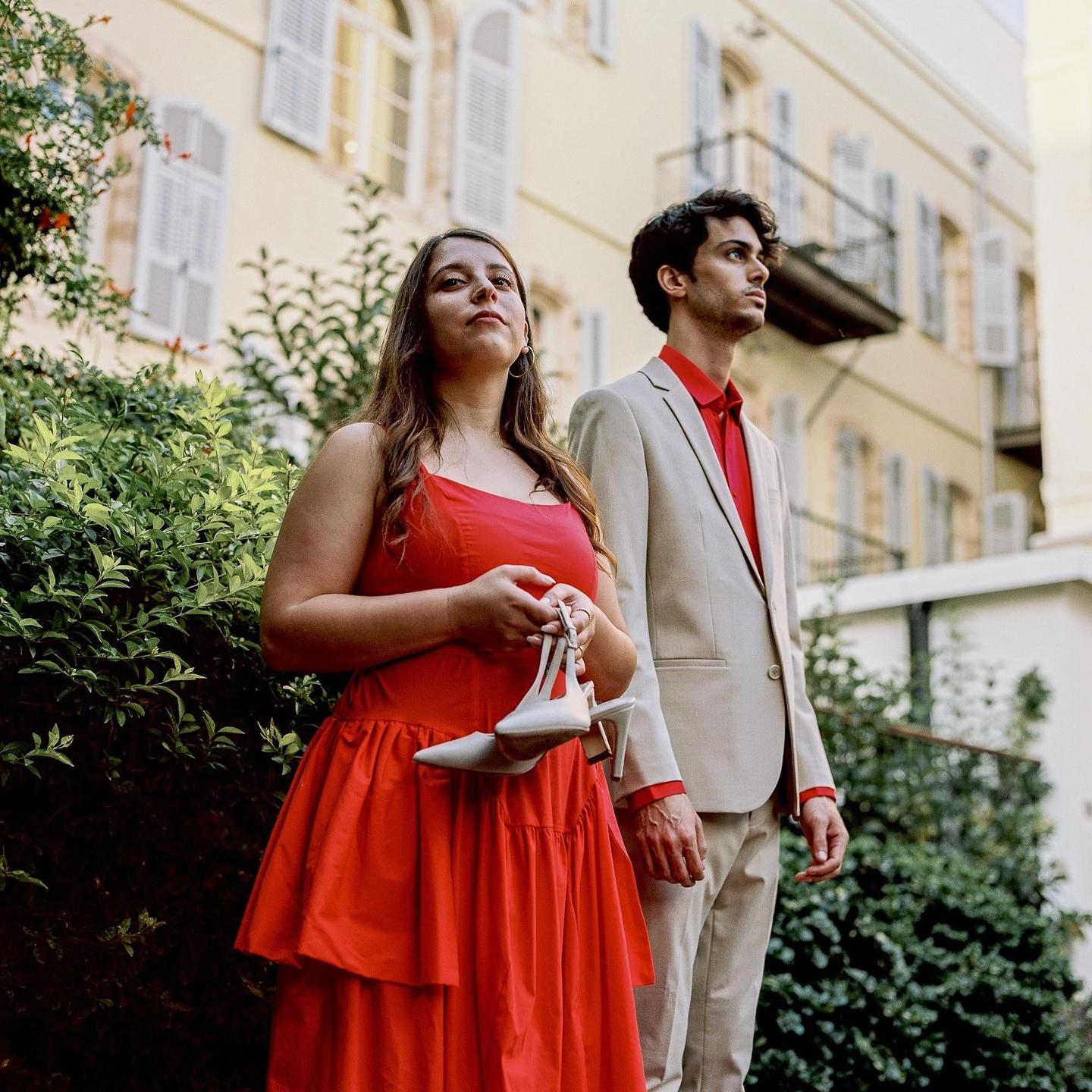A woman in a red dress holds white high heels while standing next to a man in a suit.