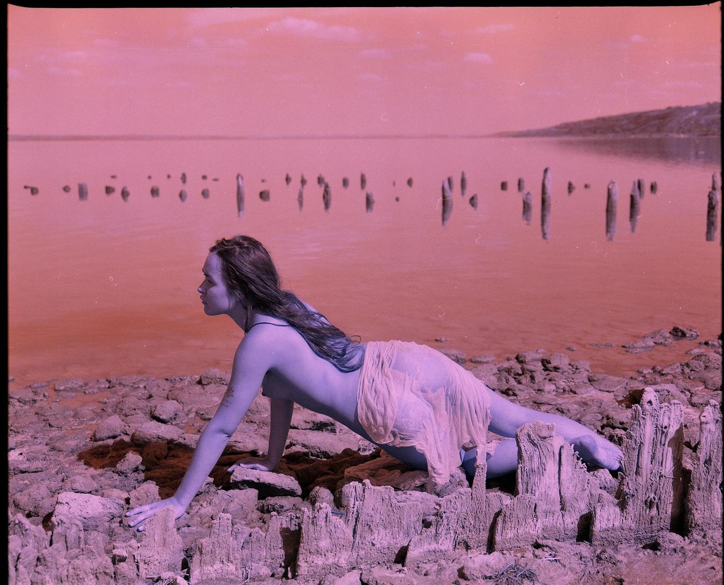 A woman with long hair crawls on a rocky shore next to a body of water with wooden posts.