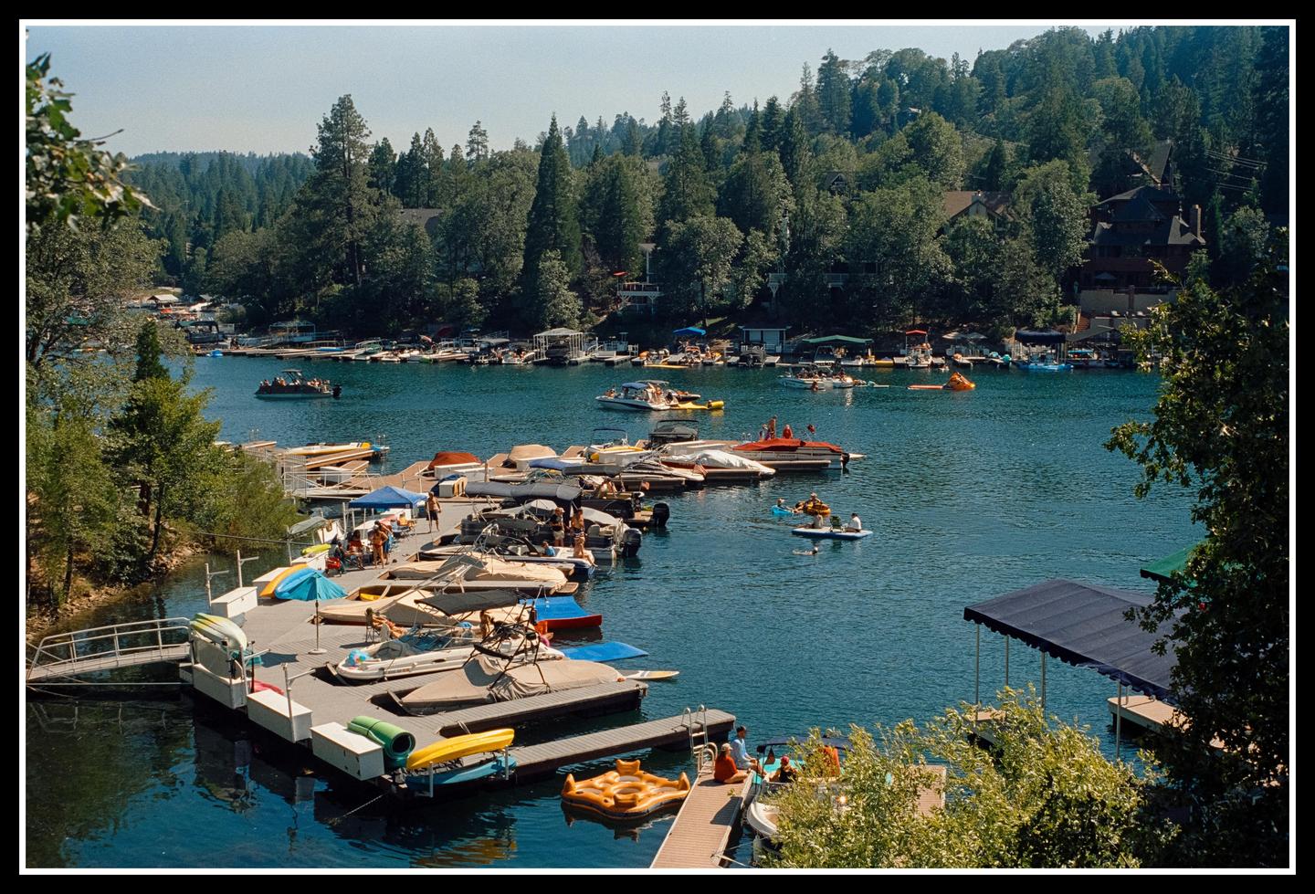 A busy lake with many boats and people on docks and in the water is surrounded by a forest and houses.