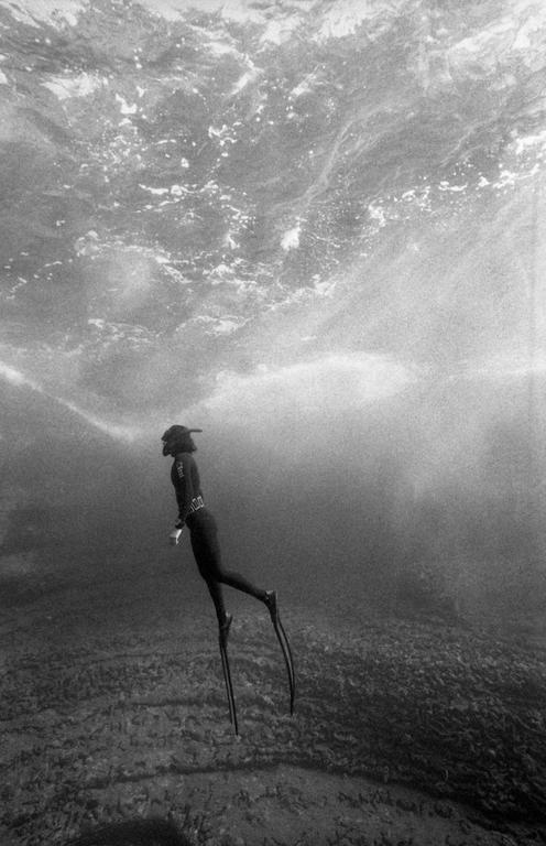 A diver in a wetsuit and fins swims underwater with light rays shining through the surface.